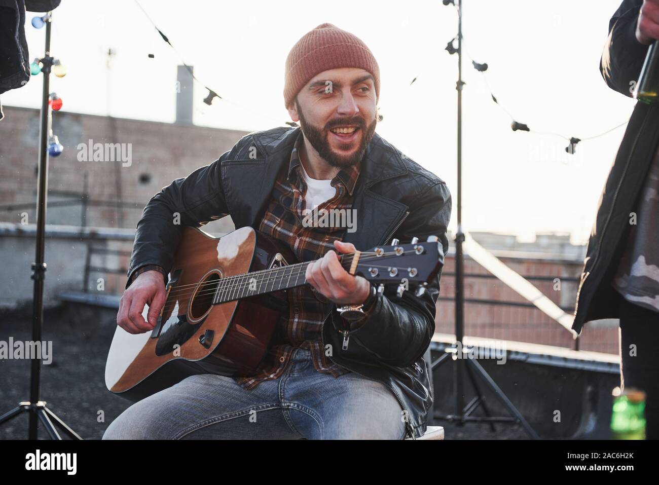 Young bearded man. Guy with acoustic guitar sings. Friends have fun at rooftop party with decorative colored light bulbs Stock Photo