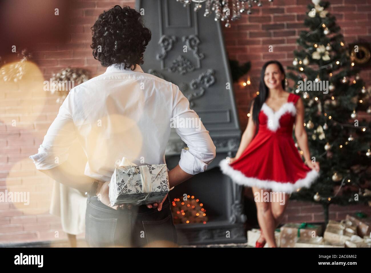 Time for sharing love and presents. Man stands and holds gift box behind. Woman in red dress will now receive Christmas gift from boyfriend Stock Photo