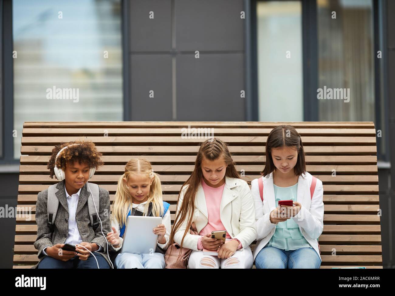 Group of school children sitting on the bench and using different gadgets they studying online Stock Photo