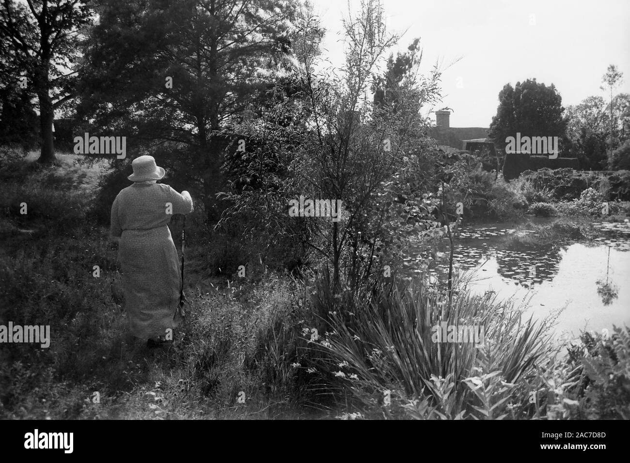 Old lady in a fine frock and hat by the Horse Pond, enjoying Christopher Lloyd's garden at Great Dixter, East Sussex.  Old black and white film photograph, circa 1980 Stock Photo