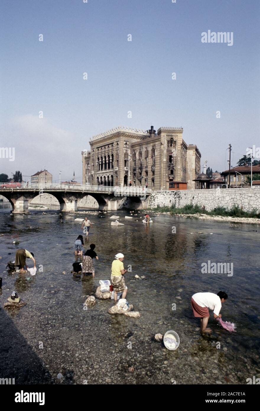 9th August 1993 During the Siege of Sarajevo: women wash laundry in the River Miljacka, with the burned-out National Art Gallery and Library in the background. Today, the Library is Sarajevo's City Hall. Stock Photo