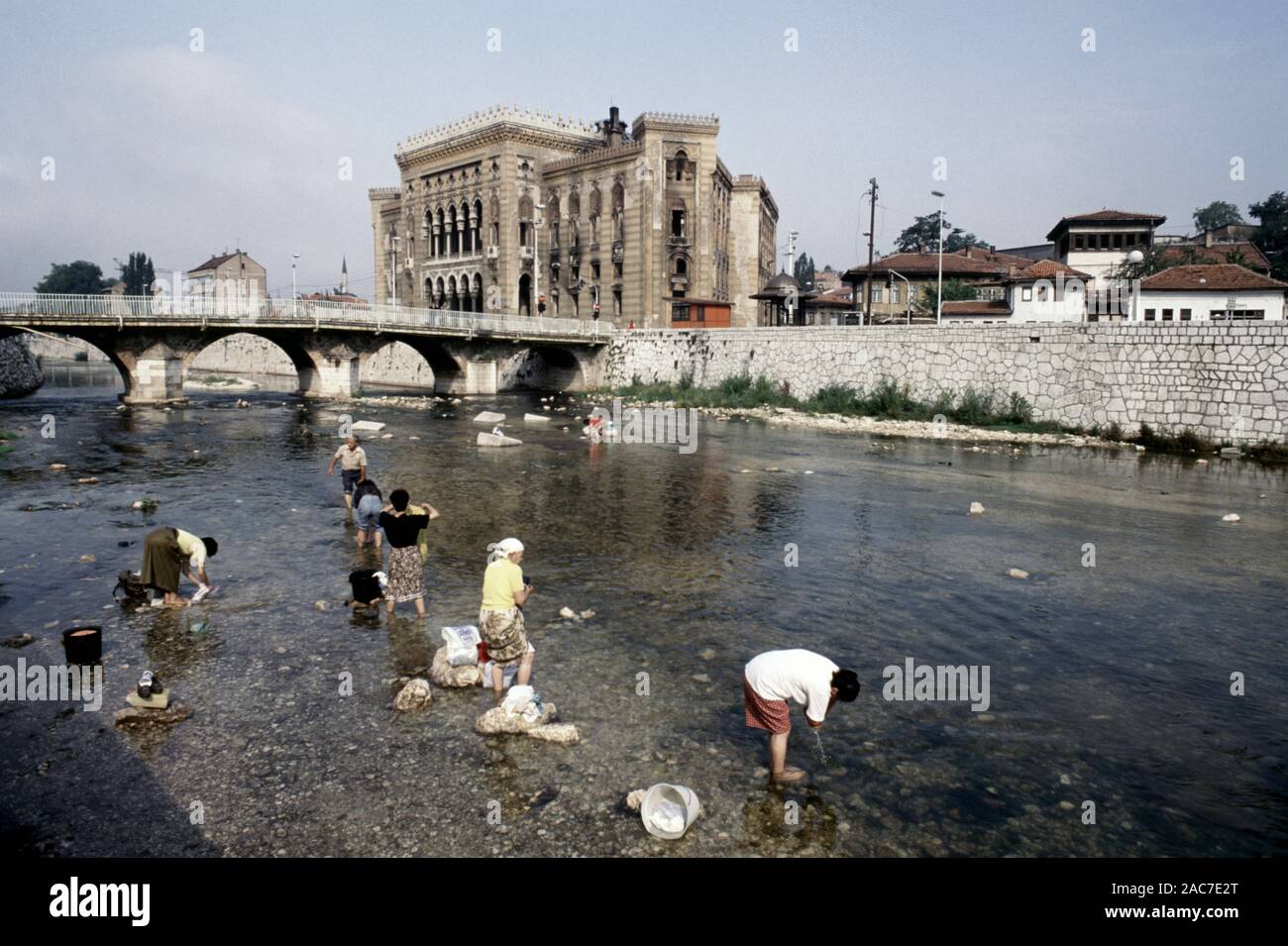 9th August 1993 During the Siege of Sarajevo: women wash laundry in the River Miljacka, with the burned-out National Art Gallery and Library in the background. Today, the Library is Sarajevo's City Hall. Stock Photo