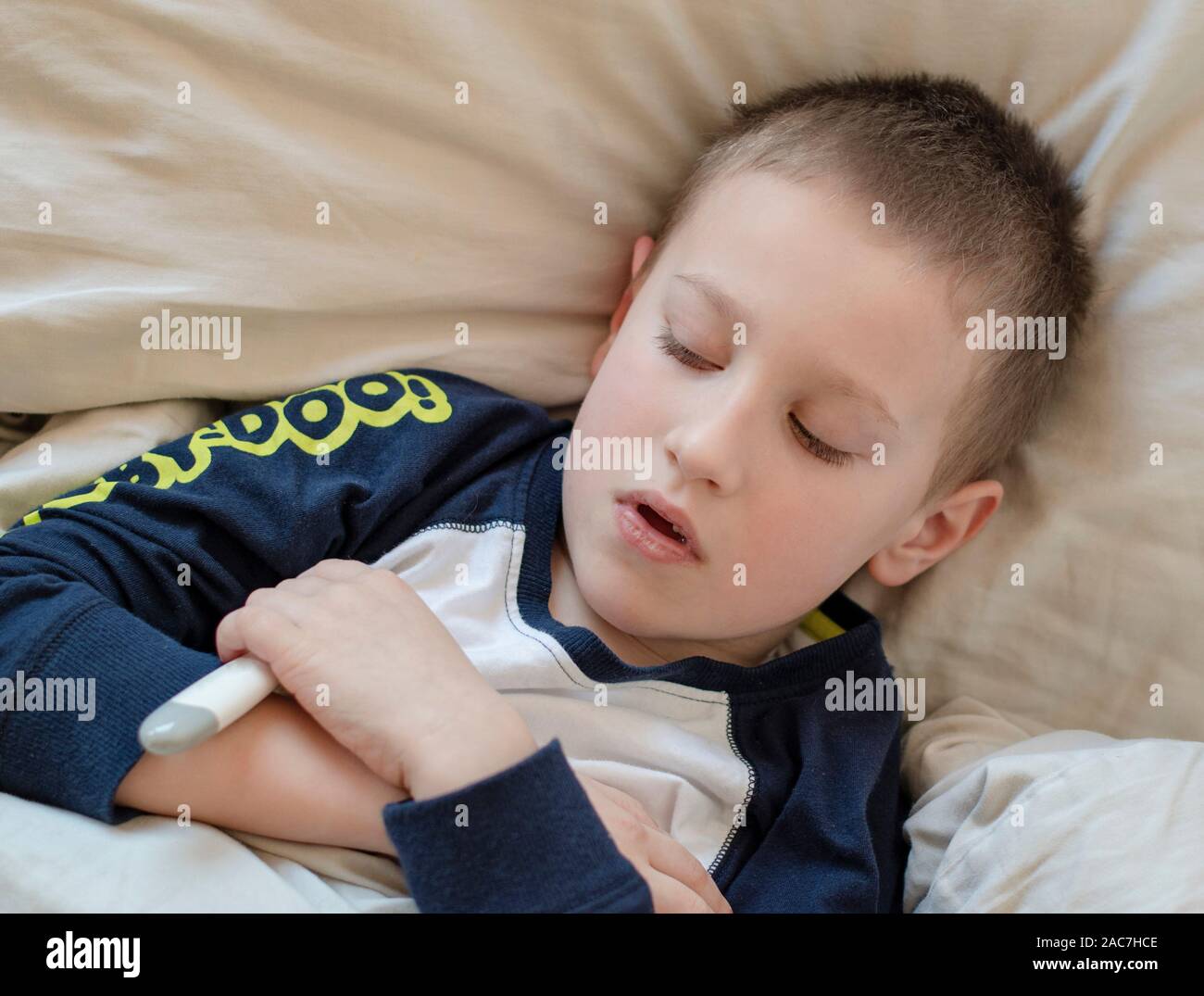 Pre-school sick boy in pyjama lying on pillow in bed, holding digital thermometer in hand. Ill boy is measuring body temperature and doesn’t feel well Stock Photo