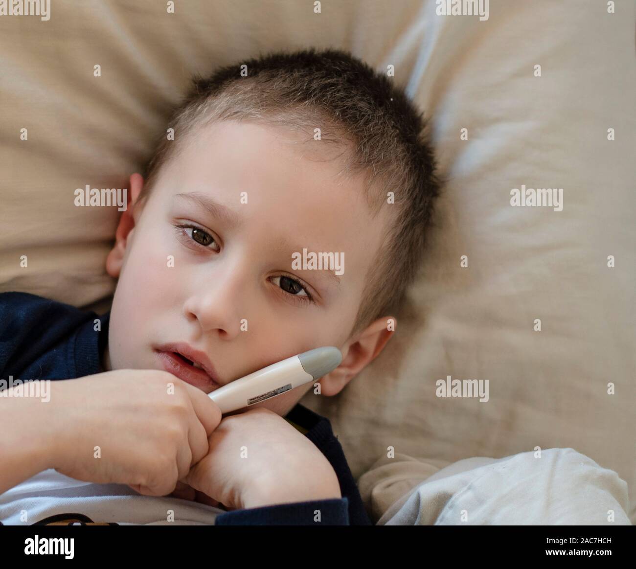 Pre-school sick boy in pyjama lying on pillow in bed, holding digital thermometer in hand and looking into camera. Ill boy is measuring body temperatu Stock Photo