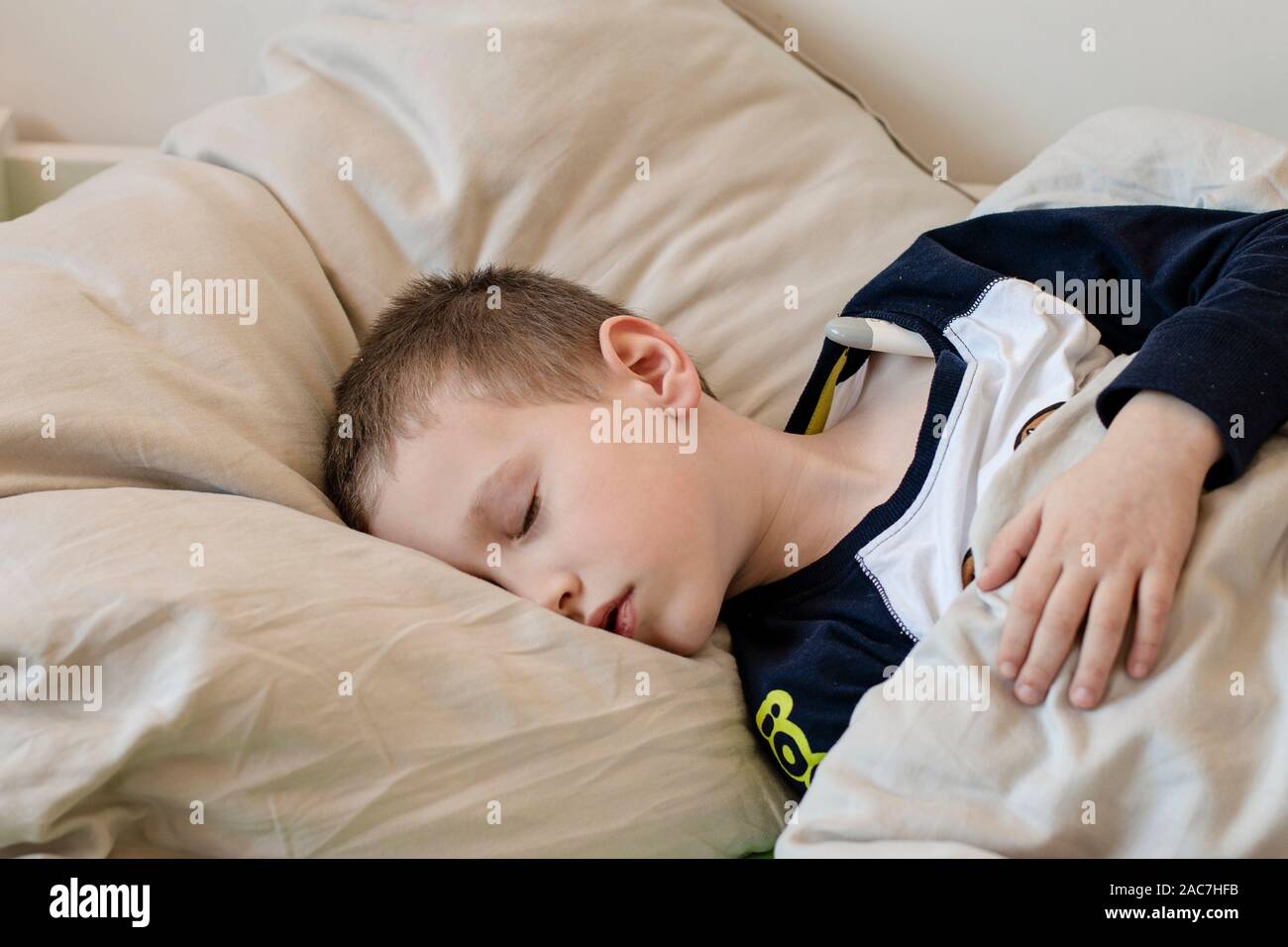 Pre-school sick boy with closed eyes lying on pillow in bed with a digital thermometer. Ill boy is measuring body temperature and doesn’t feel well. - Stock Photo