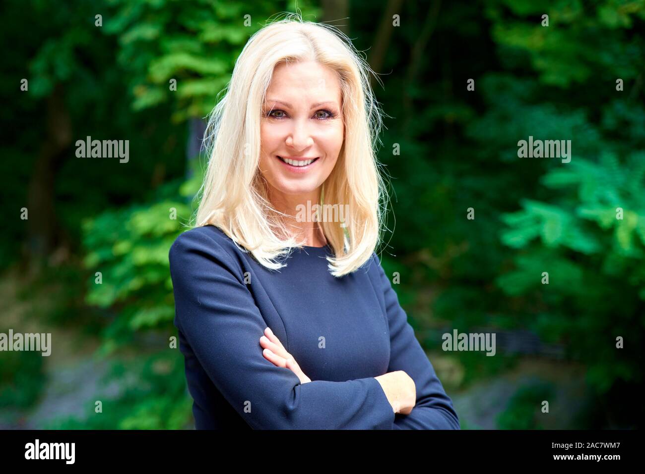 Close-up portrait shot of an elegant mature businesswoman standing with arms crossed outdoor while looking at camera and smiling. Stock Photo