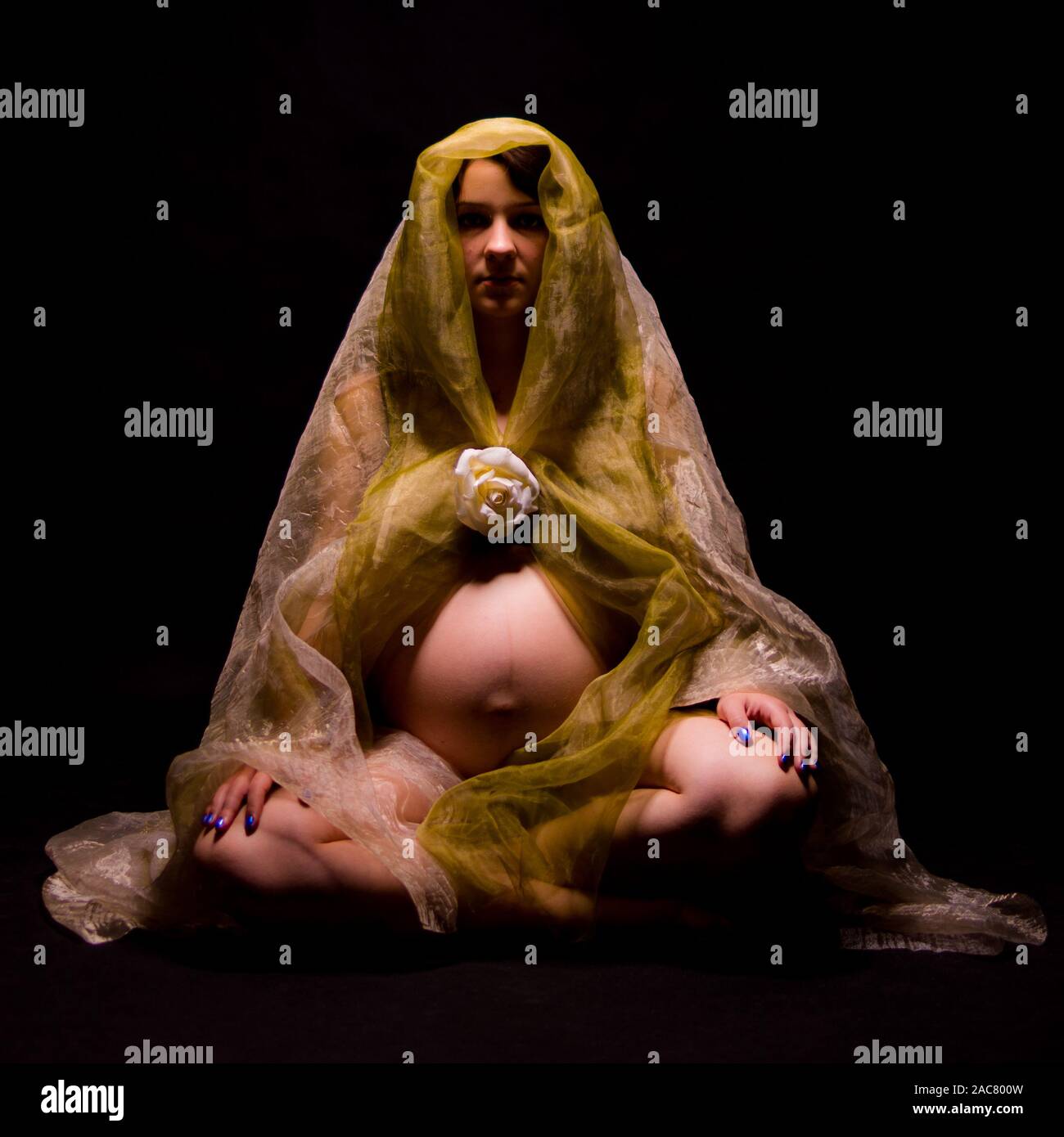 Pregnant woman sitting in the dark dressed in a transparent, delicate fabric Stock Photo