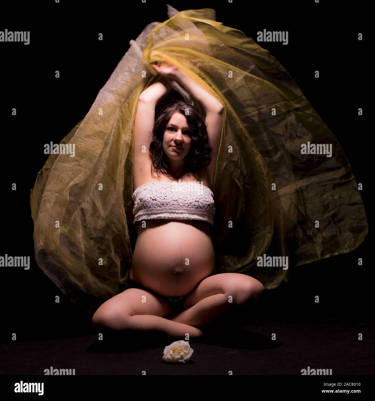 Pregnant woman dancing seated in the dark with a transparent, delicate fabric Stock Photo