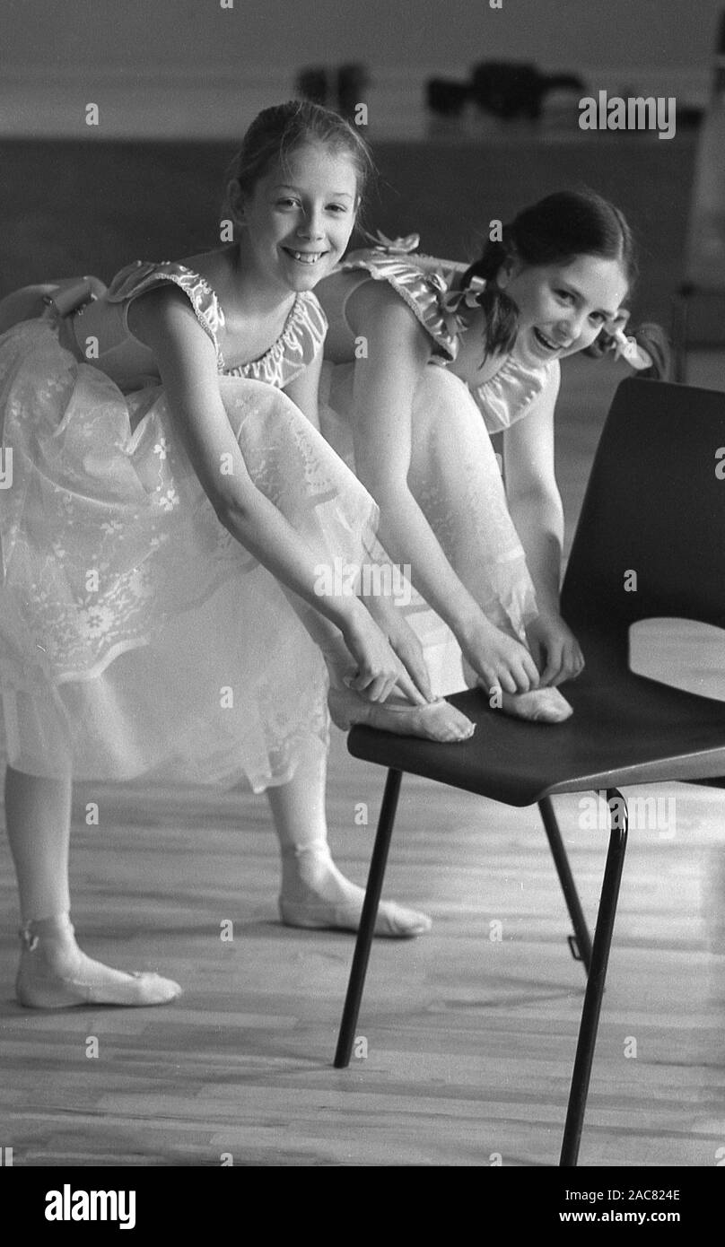 1980s, historical, two young girls in dance costumes at a ballet or dance school, with each of them with one of their ballet or pointe shoes positioned on a plastic chair, England, UK. The girls are wearing a pointe shoe, a type of shoe worn by ballet dancers when training or performing, as they enable dancers to dance on the tips of their toes for extended periods of time. Stock Photo