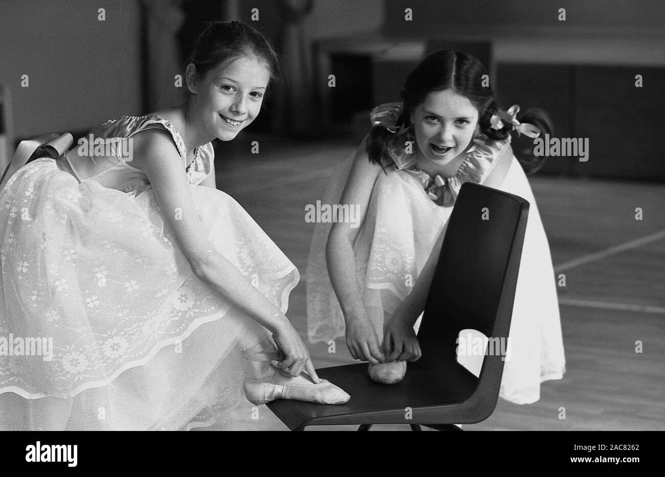 1980s, historical, 'it's all in the feet'...inside a ballet or dance school, two young girls, ballet dancers, point to their toes, England, UK. The girls are wearing a pointe shoe, a type of shoe worn by ballet dancers when training or performing, as they enable dancers to dance on the tips of their toes for extended periods of time. Stock Photo