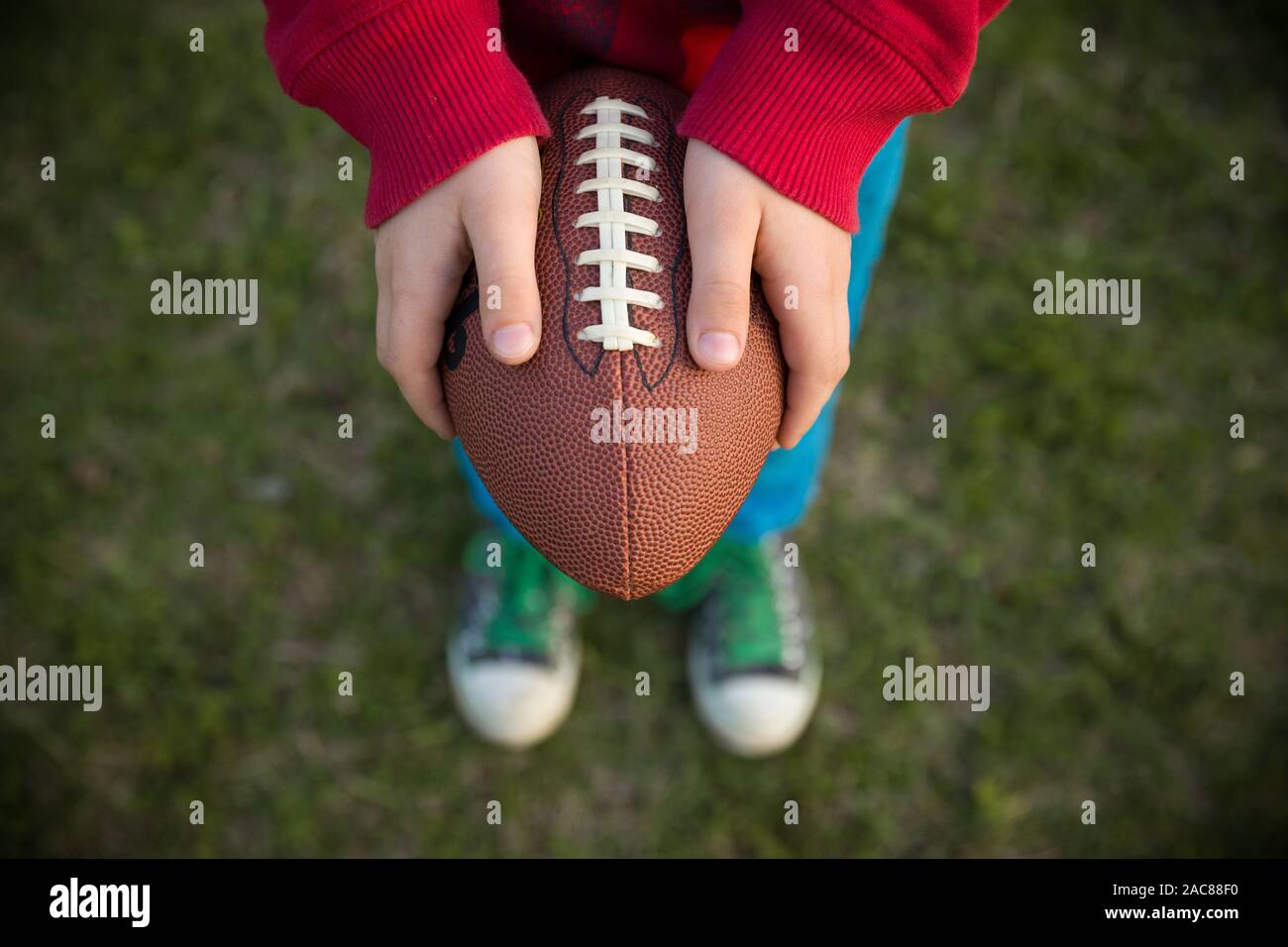 Top view on hands of little kid boy holding football on the stadium on a sunny day. Child ready to throw a football. Sport concept. Sport activities f Stock Photo