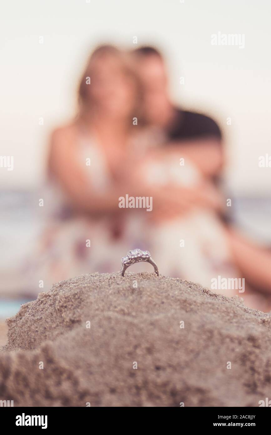 A couple sits on the beach in front of their wedding ring during an engagement shoot Stock Photo