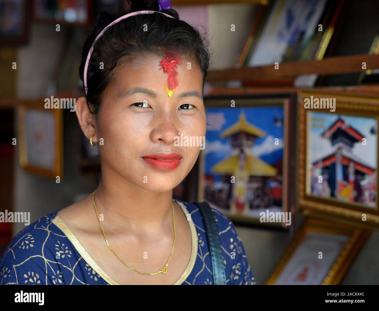 Young Nepali Hindu woman with red rice tilak mark on her forehead poses for the camera in front of souvenir photographs of Manakamana Temple. Stock Photo
