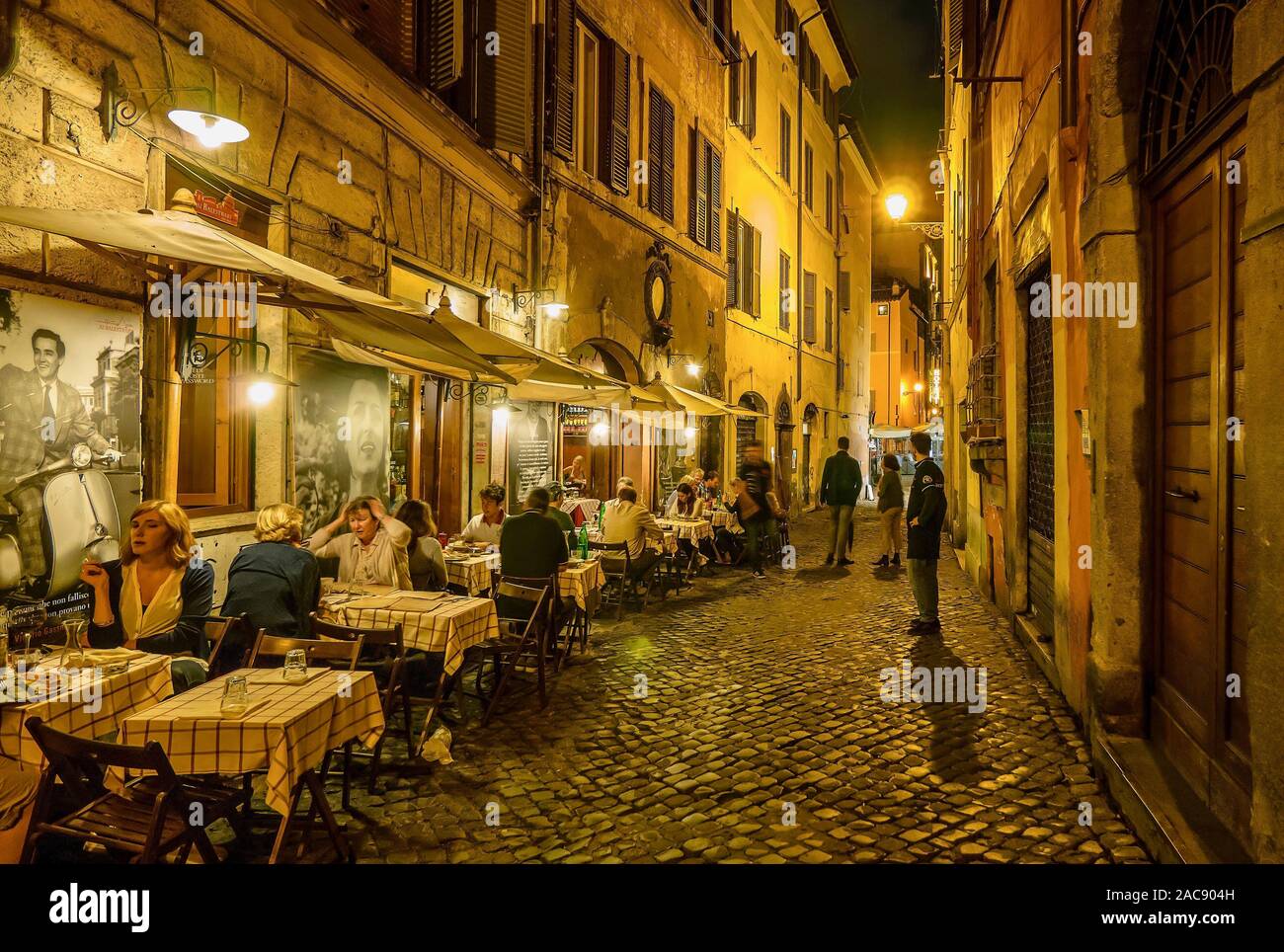 A typical residential street in Rome, where people are dining outdoors at a small neighborhood restaurant on a cobblestone street. Stock Photo