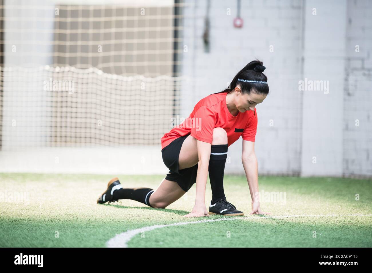 Young active female footballer in sports uniform doing exercise for stretching on the field before game Stock Photo