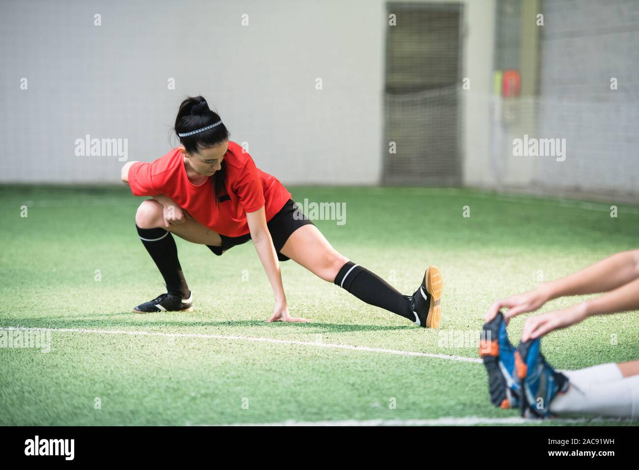 Active girl in sports uniform doing exercise for stretching legs on football field while working out before game Stock Photo