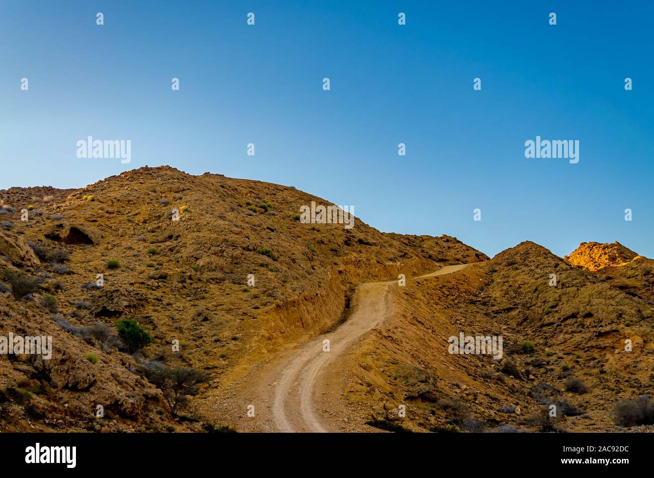 Winding dirt road through the orange colored barren mountains with a clear blue sky as background. From Muscat, Oman. Stock Photo