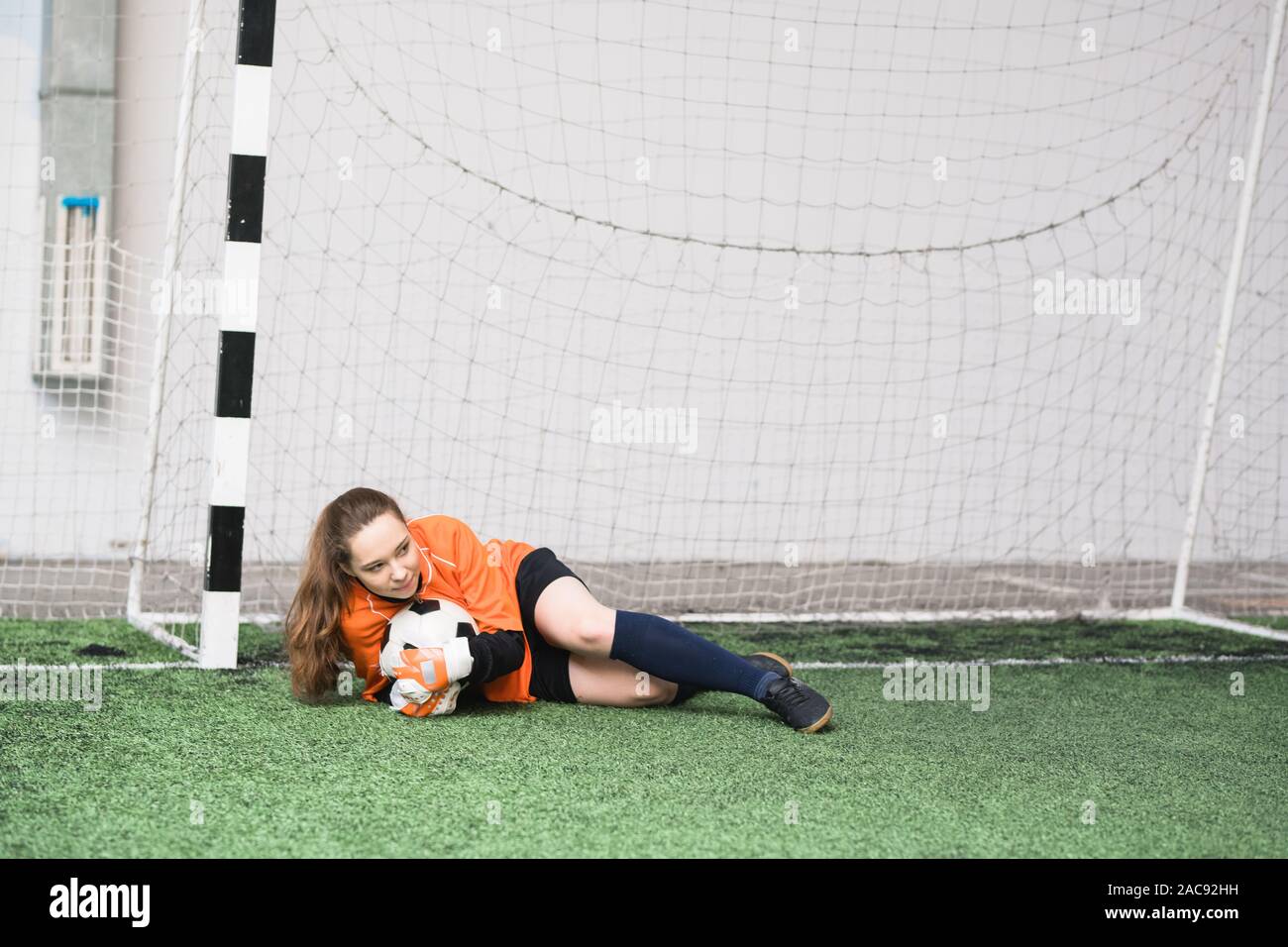 Young female goalkeeper with soccer ball lying on green field in the gates during game of football Stock Photo