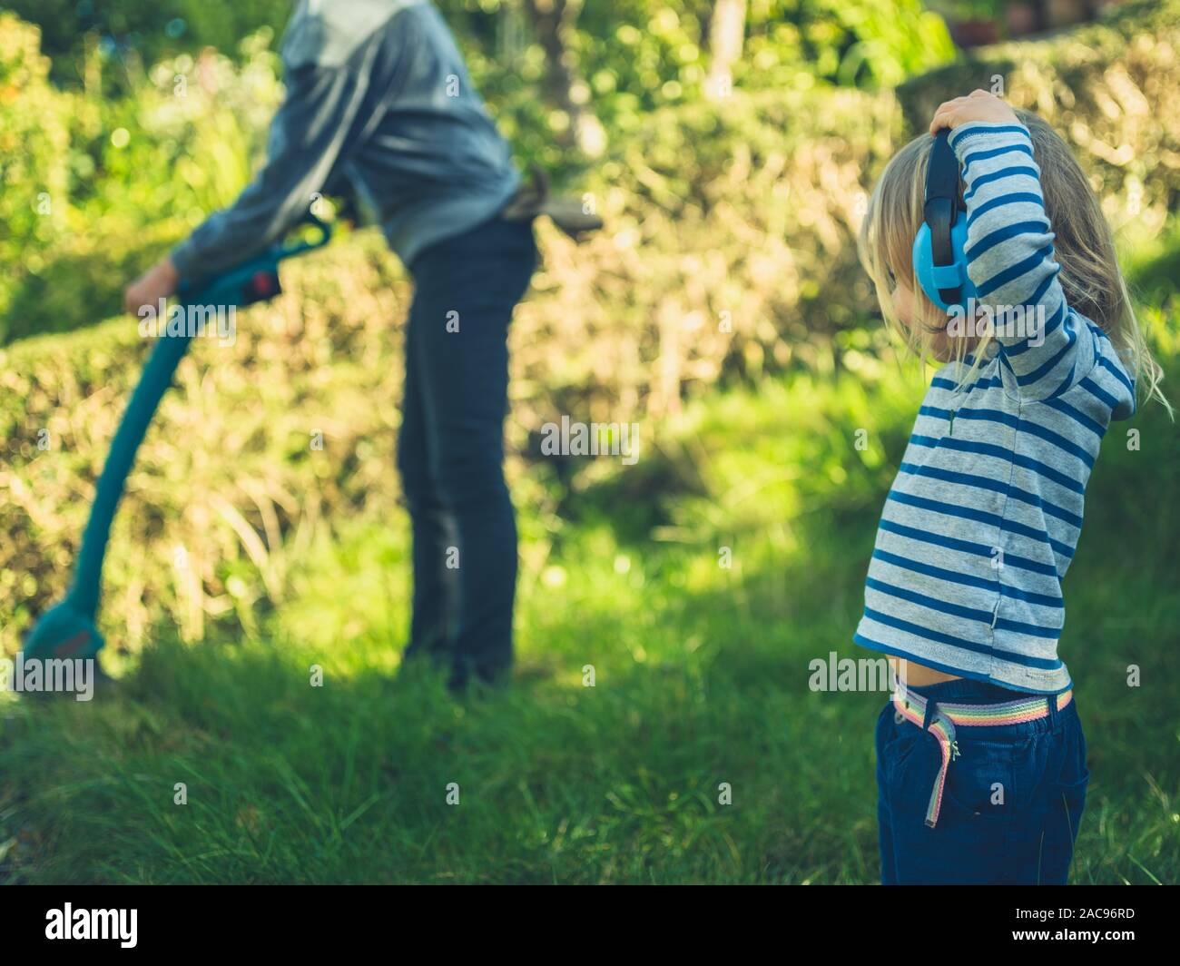 A little toddler wearing ear defenders is watching his mother strim the grass in their garden Stock Photo