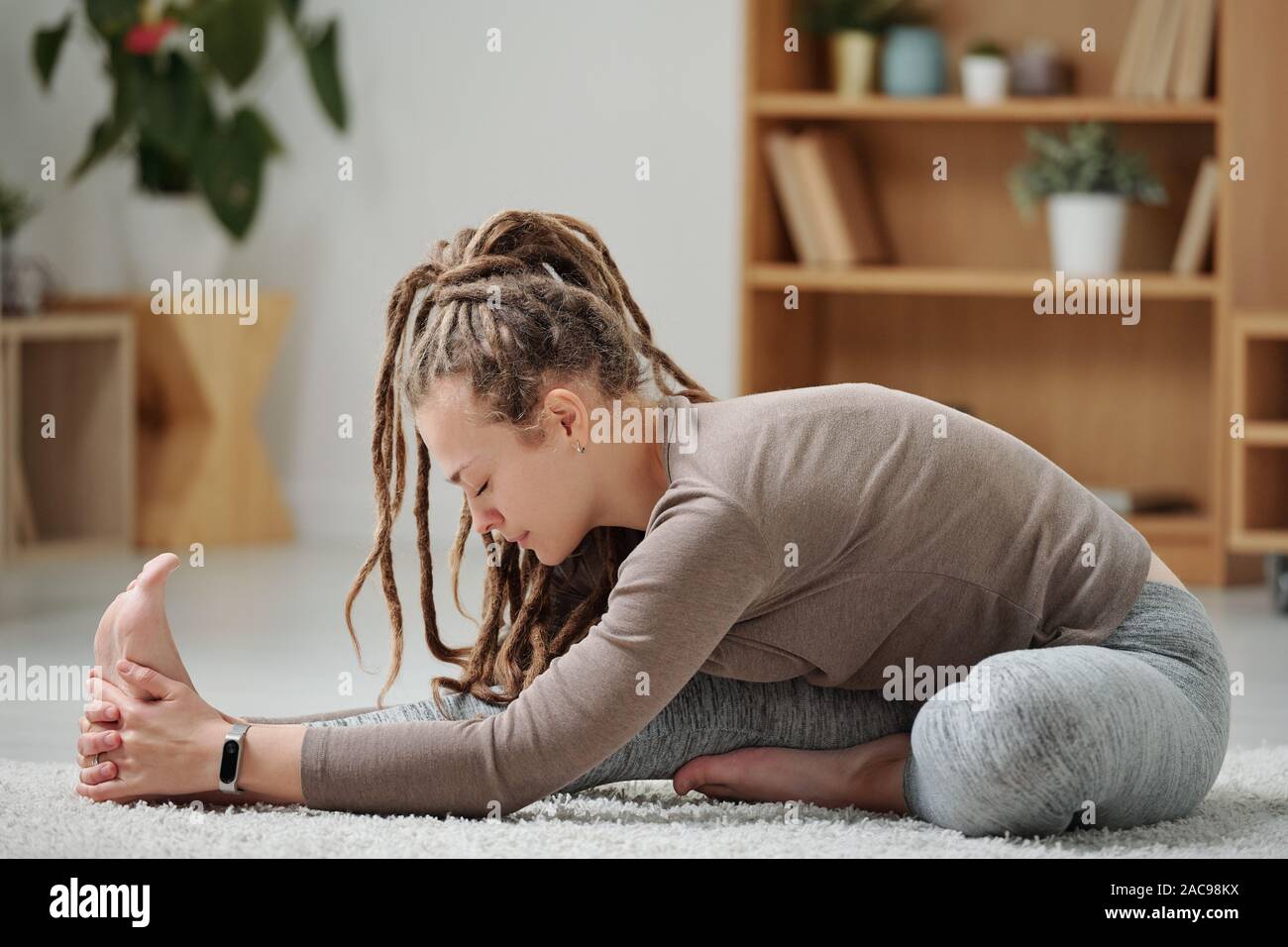 Young flexible woman in activewear sitting on the floor and stretching one leg Stock Photo
