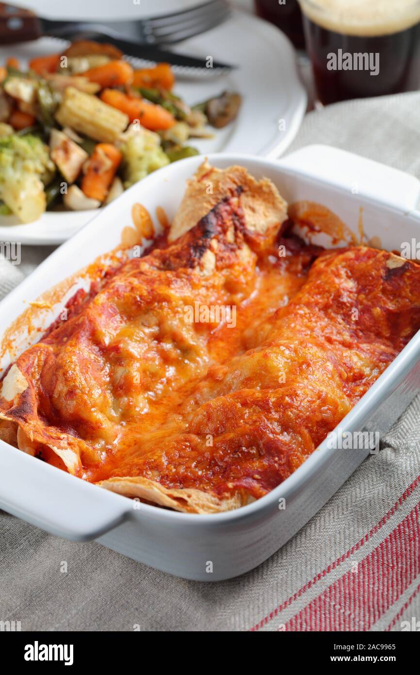 Two enchiladas in a baking dish with a portion of roasted vegetables and a glass of beer Stock Photo