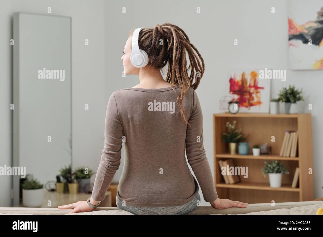Back view of young restful female in activewear and headphones sitting on sofa Stock Photo