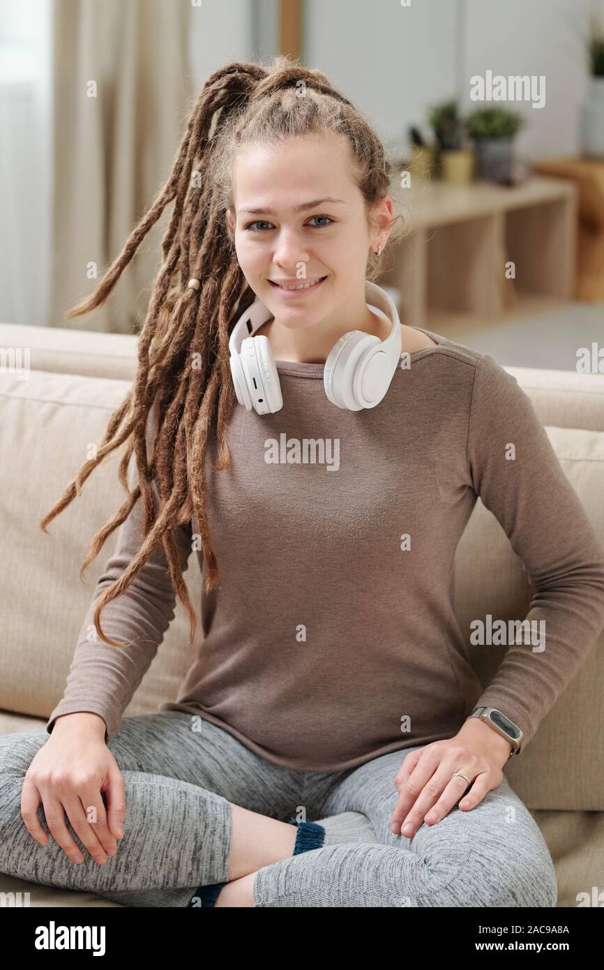 Pretty smiling girl in activewear and headphones sitting on couch at home Stock Photo
