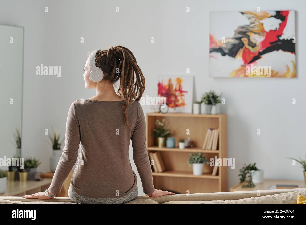 Back view of restful girl with dreadlocks sitting on sofa and listening to music Stock Photo