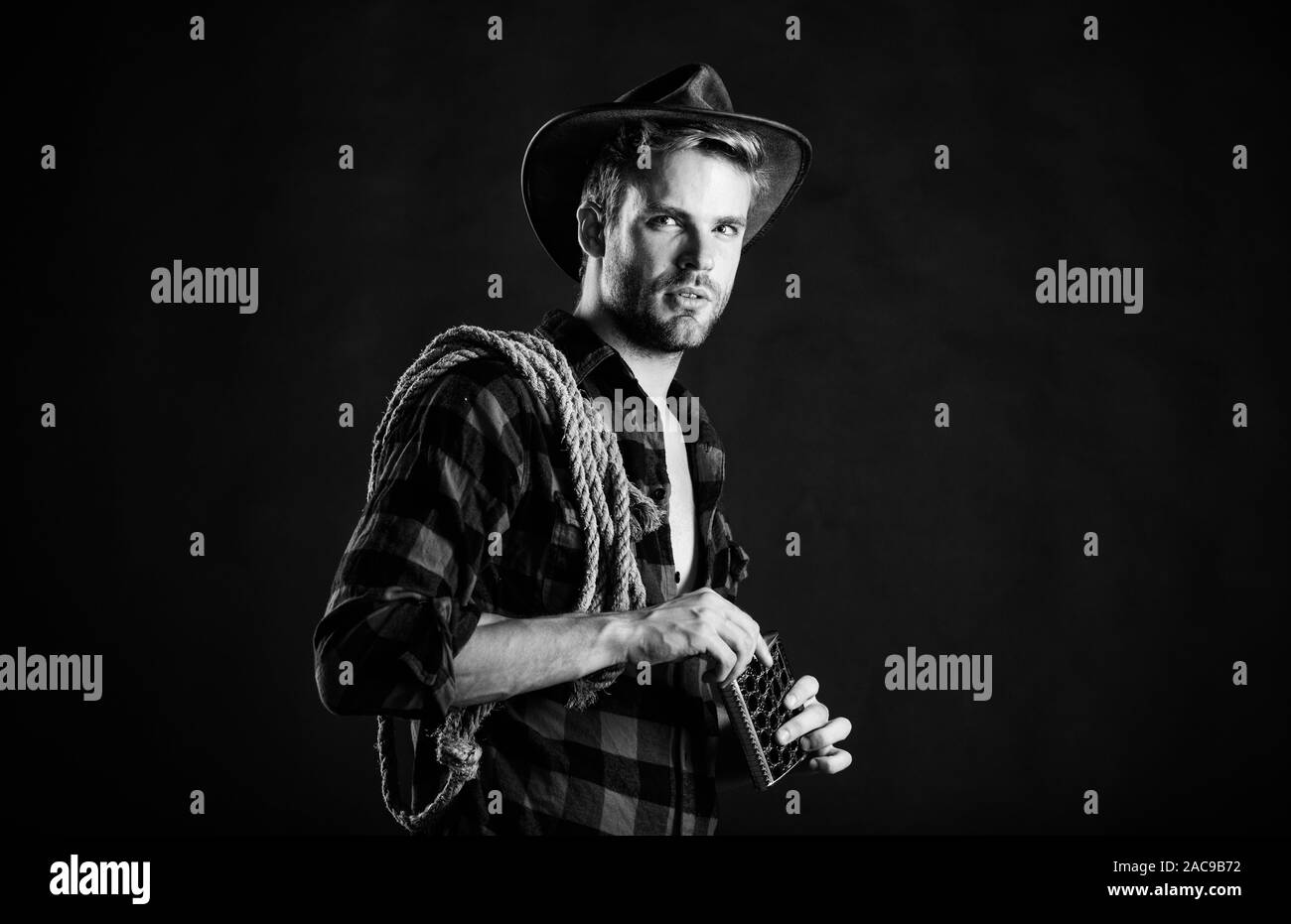 Western culture. Man handsome unshaven cowboy black background. Western life. Man wearing hat hold rope and flask. Lasso tool of American cowboy. Sher Stock Photo