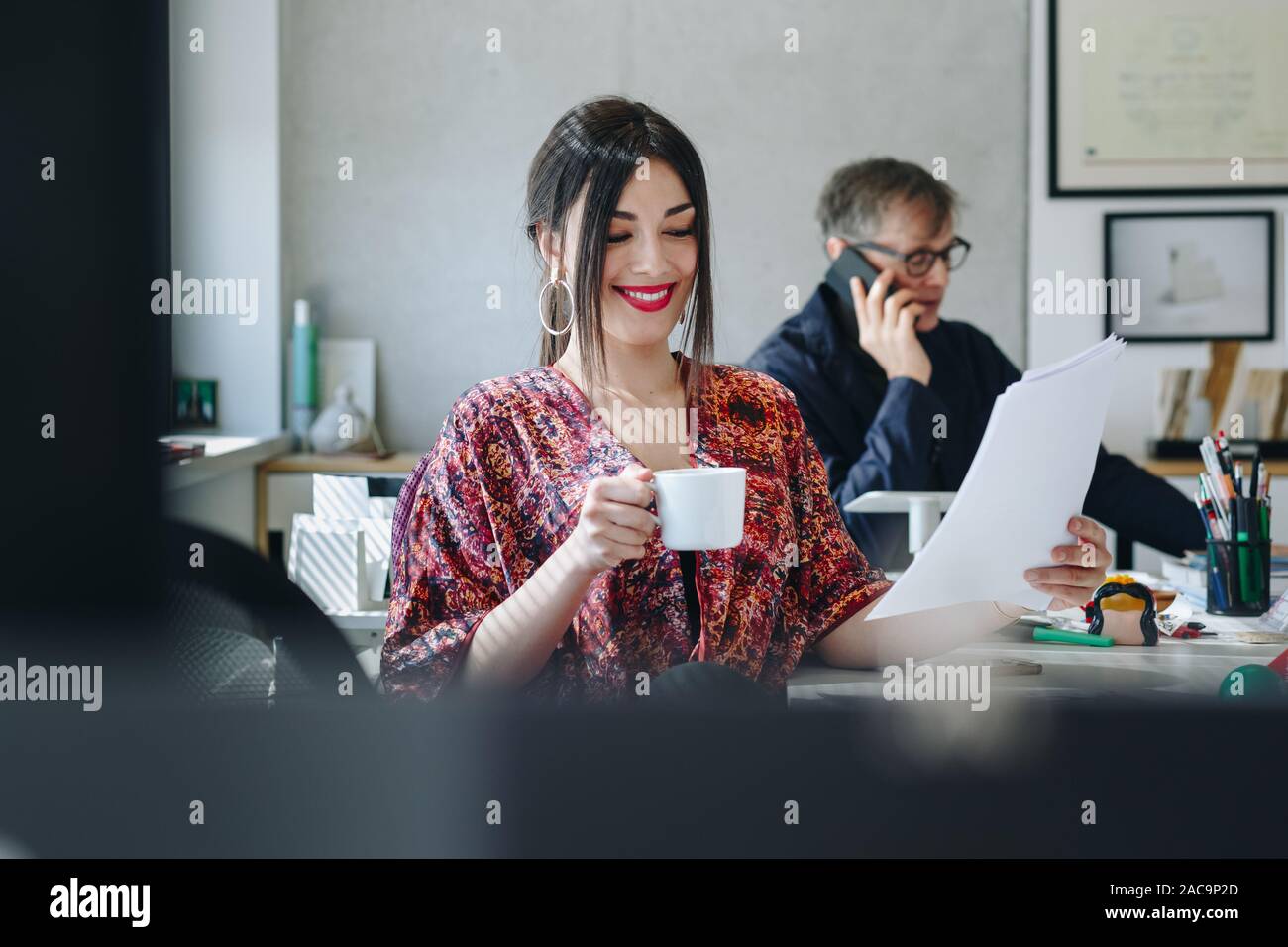 Portrait of  happy businesswoman drinking coffee in creative office. Coworker talking on the phone in the background. Happy and busy office life conce Stock Photo