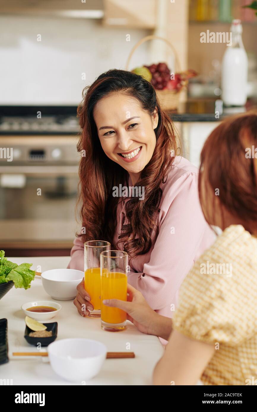Happy mature Vietnamese woman enjoying orange juice when talking to daughter at breakfast Stock Photo