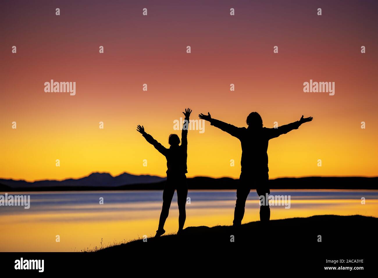Silhouettes of two hikers stands in winner pose with raised arms against sunset lake and mountains Stock Photo