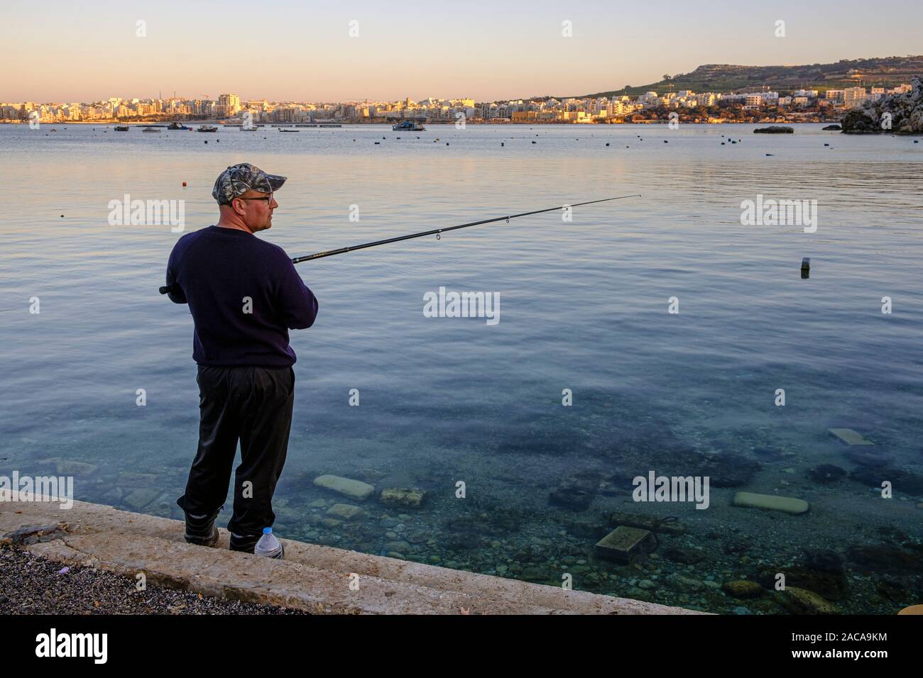 Fishing in the evening at Mistra Bay with a view across Saint Paul's Bay towards Bugibba, Malta Stock Photo