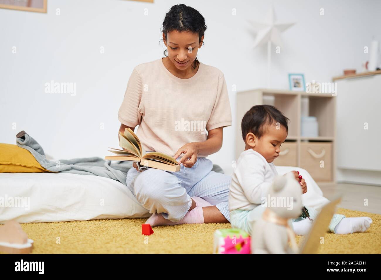 Young mother sitting on the floor and reading a book to her little ...