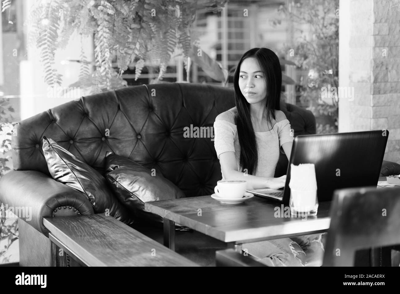 Young beautiful Asian teenage girl sitting while thinking at outdoor coffee shop with laptop on wooden table Stock Photo