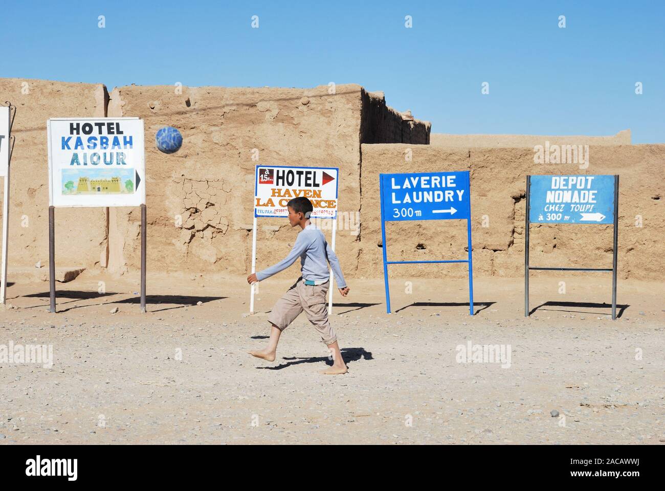 Boy playing football in a village in the desert, behind him signs to Hotel Kasbahs, Hassi Labid, South Morocco, Africa Stock Photo