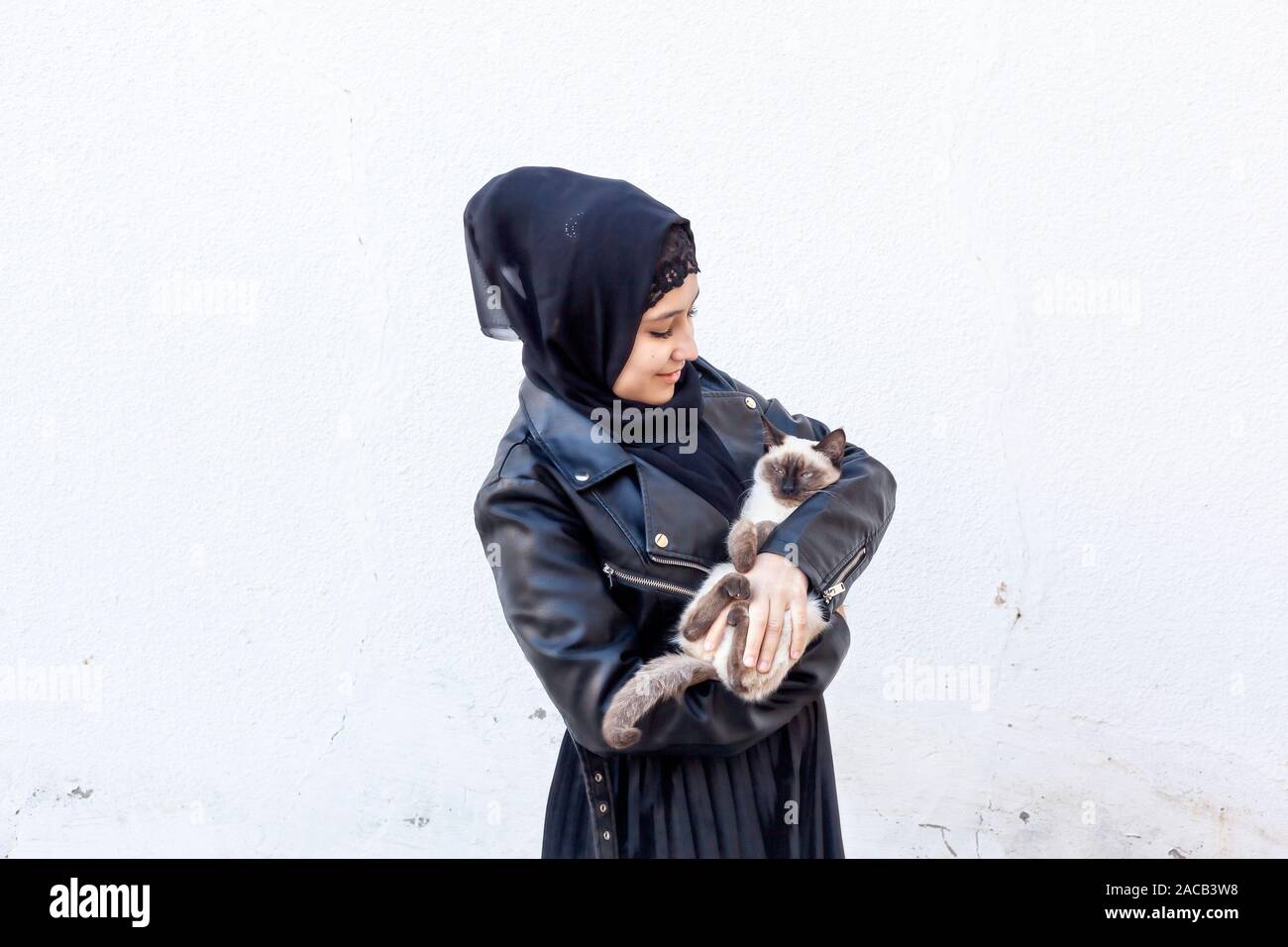 Beautiful middle-eastern Muslim woman plays with her exotic domestic Siamese cat outdoors. Cute Arabian girl in hijab and her cute pet. Pet-loving con Stock Photo