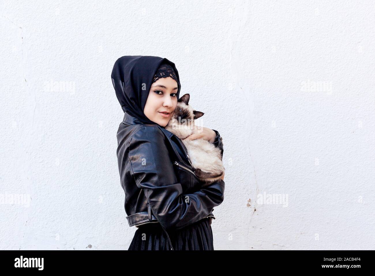 Beautiful middle-eastern Muslim woman plays with her exotic domestic Siamese cat outdoors. Cute Arabian girl in hijab and her cute pet. Pet-loving con Stock Photo