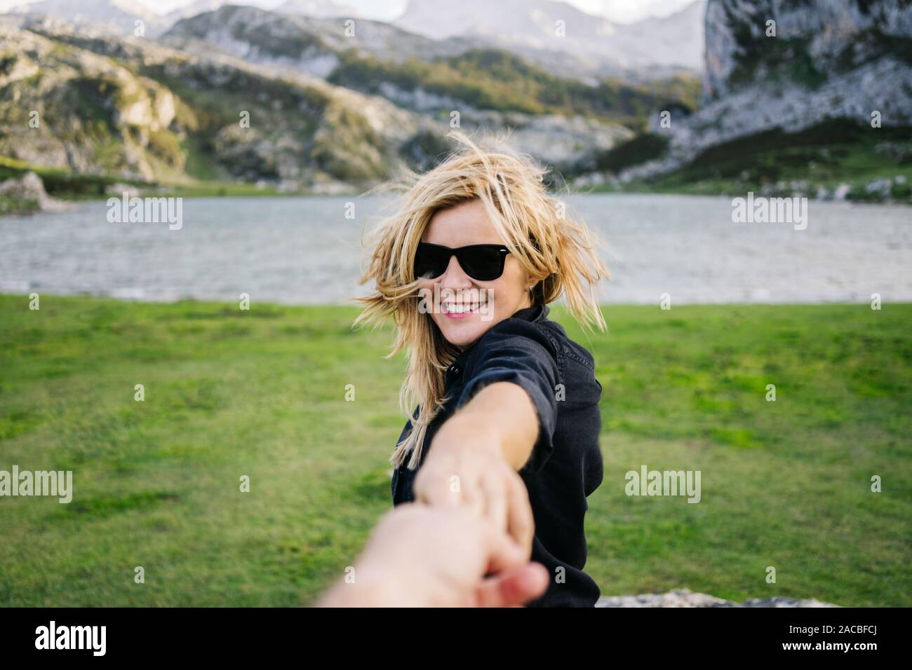 A beautiful caucasian blonde woman holds a man's hand in a mountainous landscape with lake Stock Photo