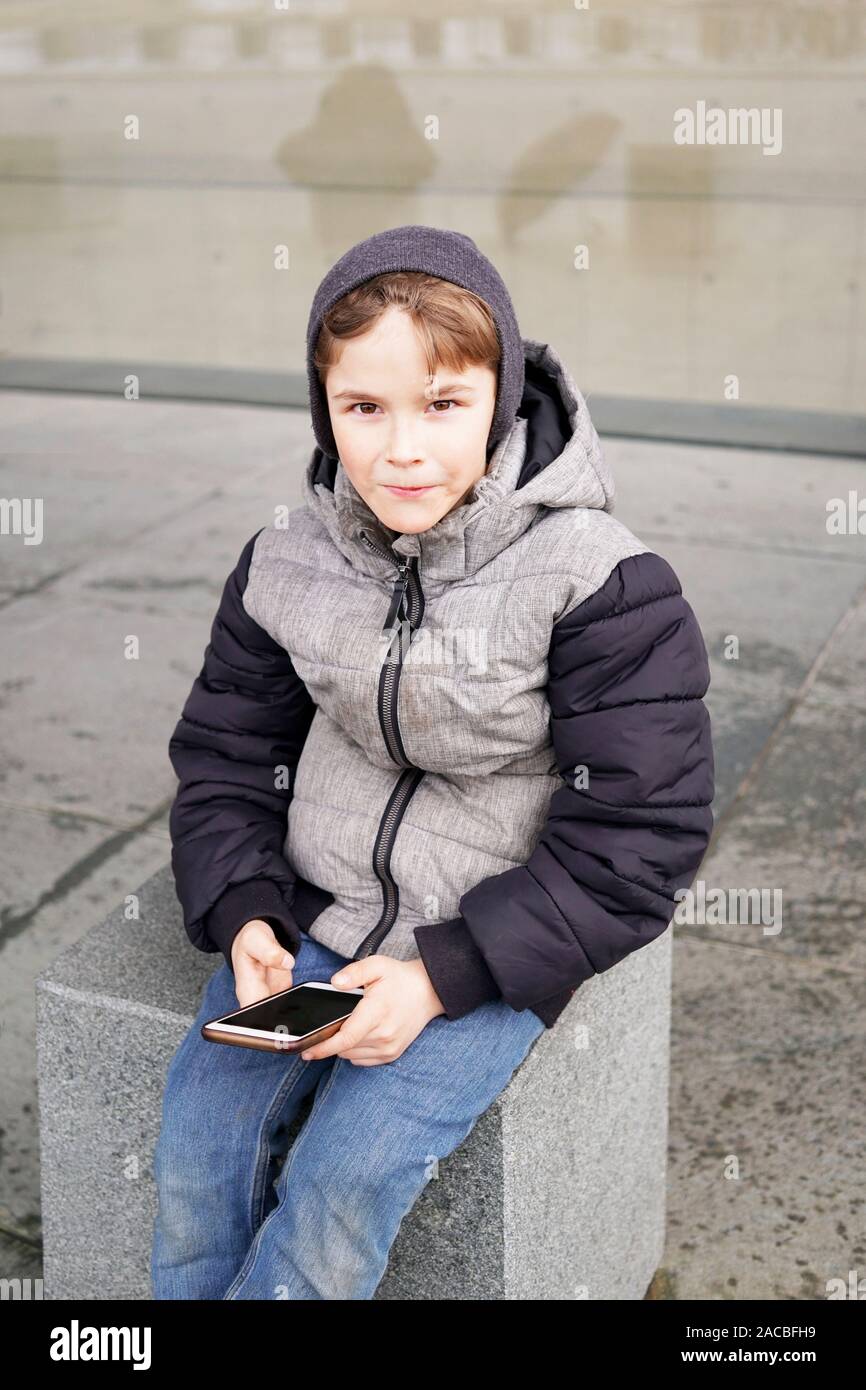 7 year old boy with smartphone mobile cell phone sitting outside alone on city street in winter Stock Photo