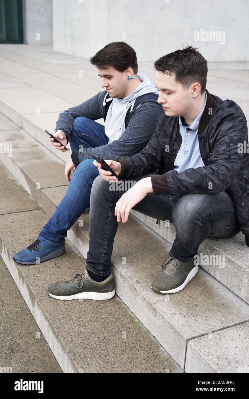 two antisocial mobile phone addicted male teenagers looking at smartphone while sitting on outside starircase - technology concept Stock Photo