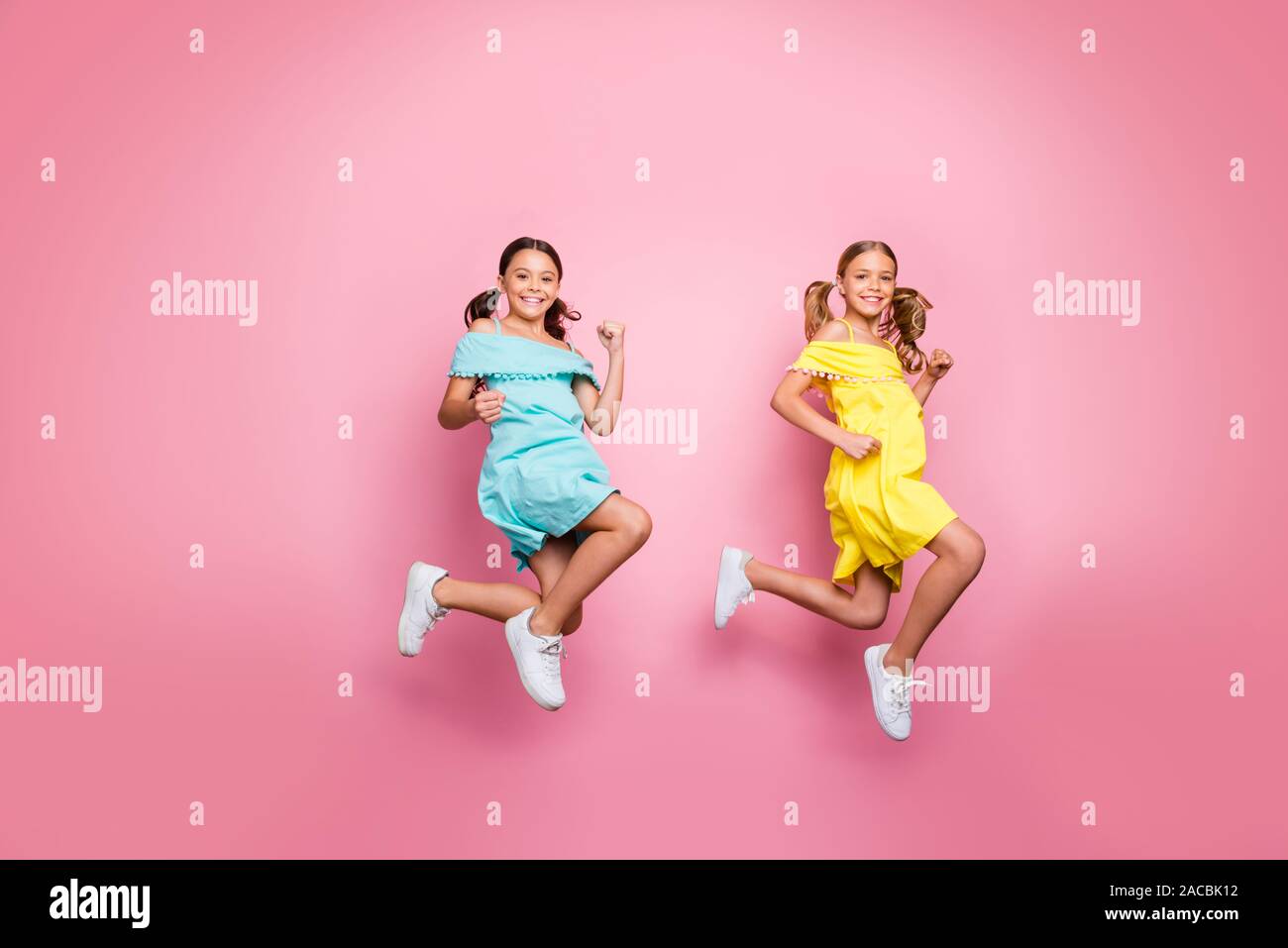 Full size profile photo of two friendship small sisters ladies models jumping high hurry home summer holidays wear blue yellow dresses isolated pink Stock Photo
