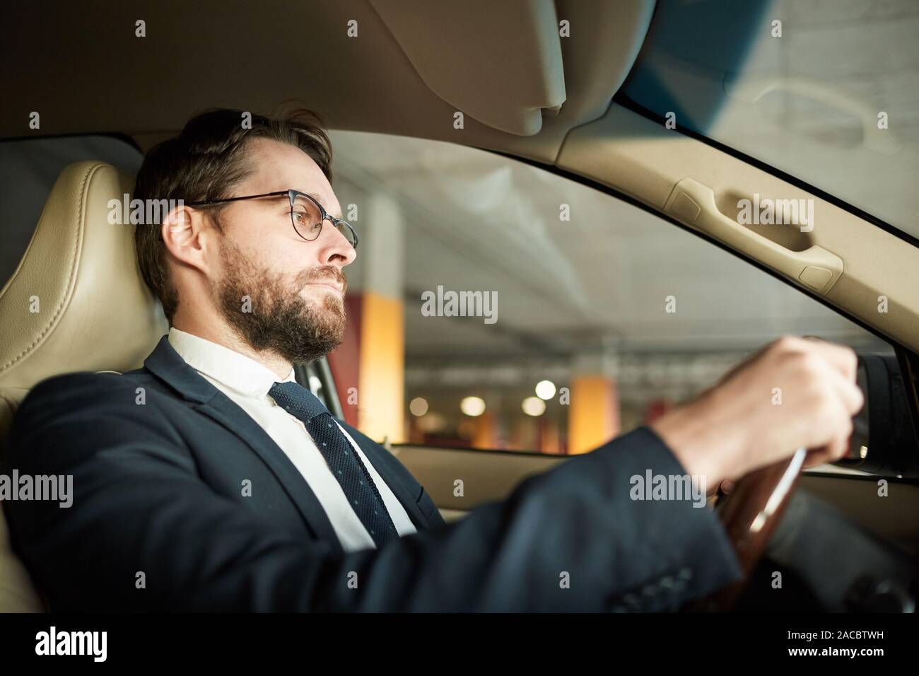 Young serious driver in eyeglasses and in suit holding the wheel and ...