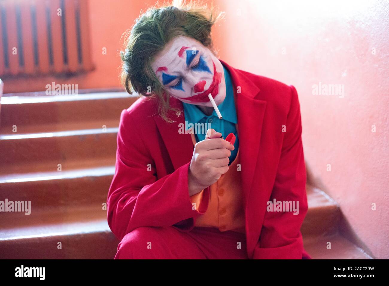 A man with a clown makeup and in a red suit sits on the stairs and smokes a  cigarette Stock Photo - Alamy