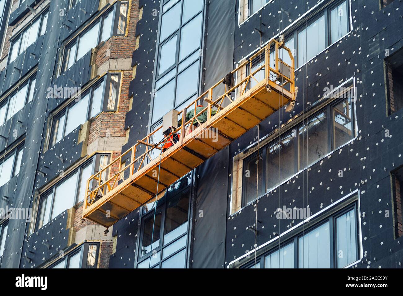 Suspended construction craddle near wall of hightower residentaial building with insulation and ventilated facade on construction site. Engineering Stock Photo