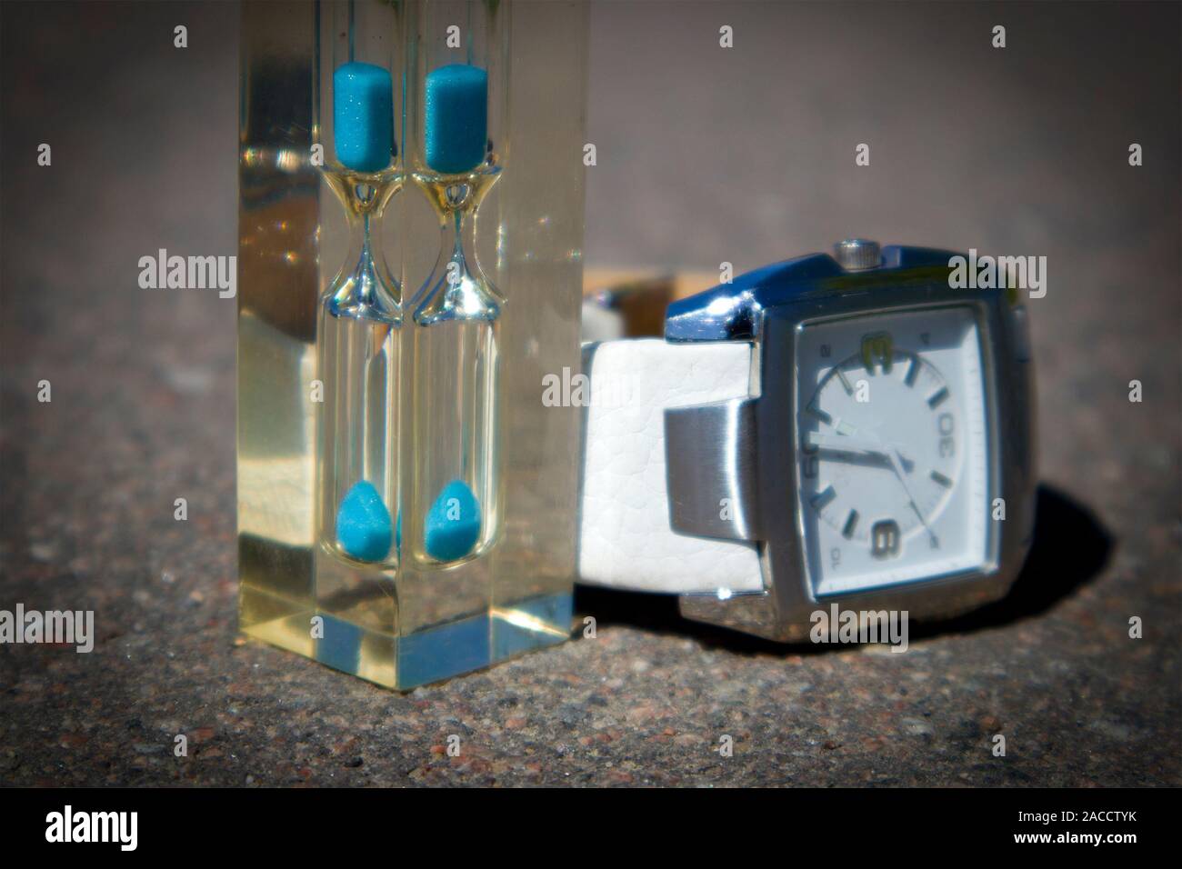 Transparent hourglass with blue sand and watch with white clock face against asphalt, time concept Stock Photo