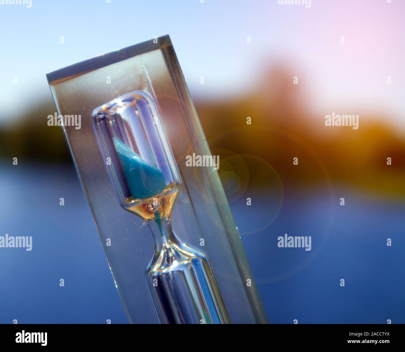 Transparent hourglass with blue sand against the landscape with calm lake, time concept Stock Photo