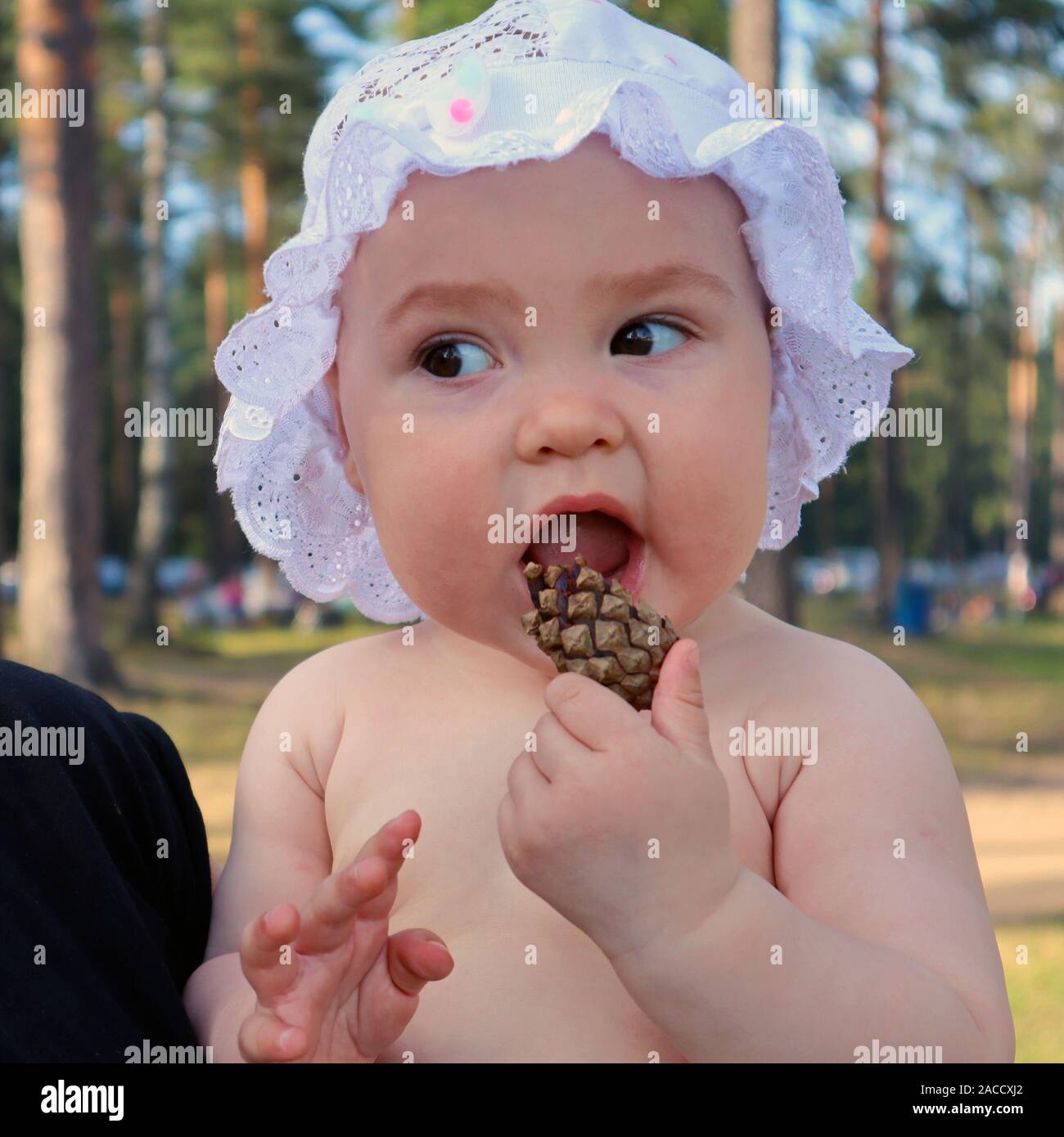 Adorable baby girl dressed in a cap with ruffles playing with pine cone outdoors Stock Photo