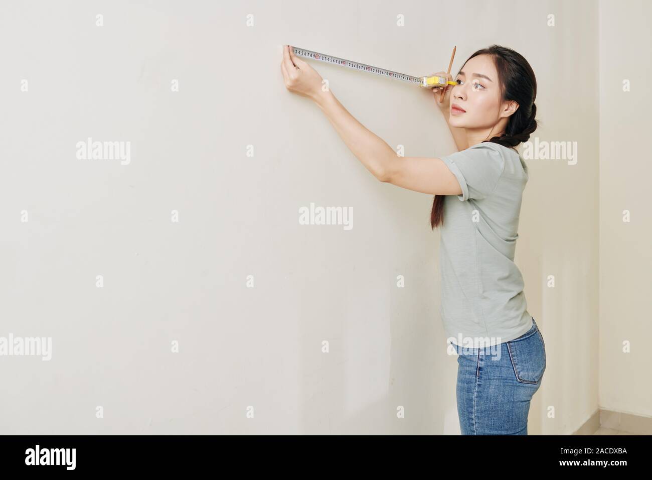 Beautiful serious young woman using tape measure when measuring wall and drawing marks Stock Photo