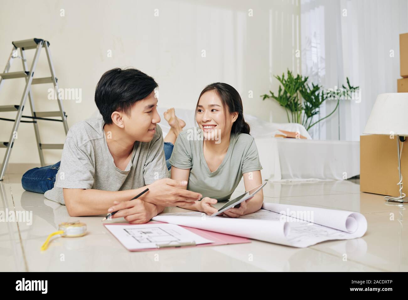 Beautiful young Vietnamese couple with tablet computer lying on the floor and discussing apartment plan Stock Photo